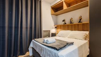 A neatly made bed with a white comforter and a folded towel, wooden shelves above the bed, and a nightstand with a lamp.