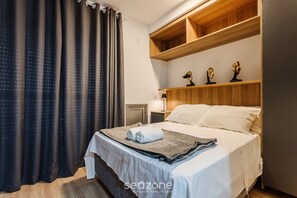 Un lit bien fait, avec une couette blanche et une serviette pliée, des étagères en bois au-dessus du lit et une table de chevet avec une lampe.
