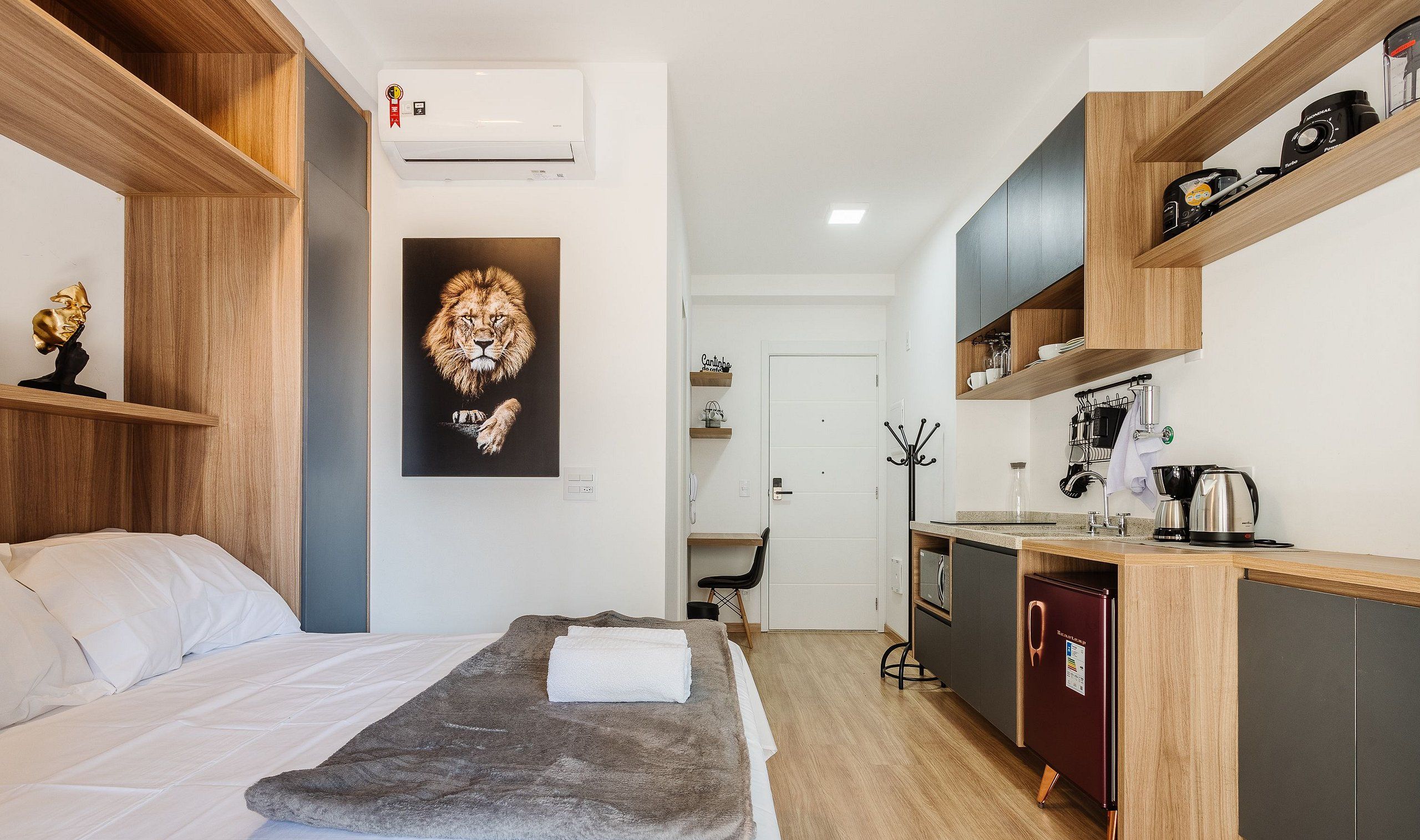 A compact hotel room with a bed, a gray throw blanket, a lion artwork, a kitchenette with a coffee maker, and a refrigerator.