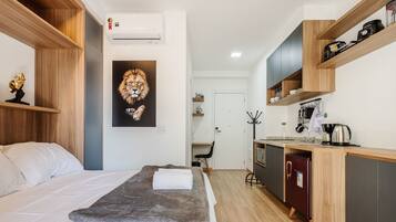 A compact hotel room with a bed, a gray throw blanket, a lion artwork, a kitchenette with a coffee maker, and a refrigerator.