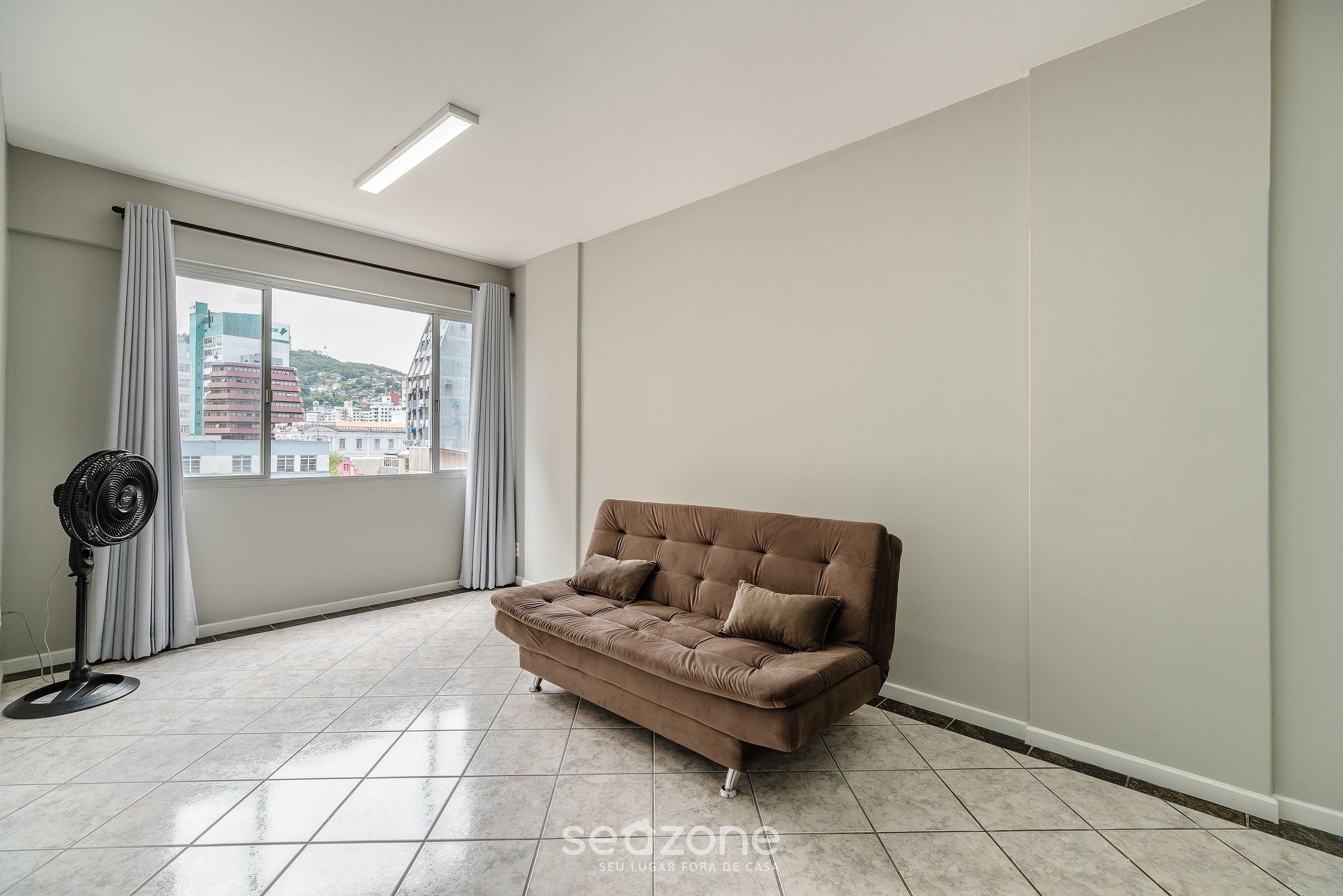 Een ruime kamer met een groot raam, een bruine bank en een ventilatorkachel.
