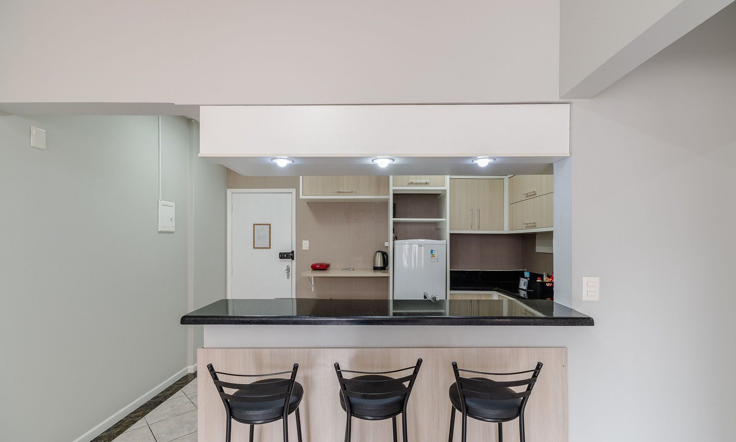 A compact kitchen with a black countertop, three black chairs, and a refrigerator.