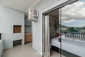 A modern bedroom with a bed, a sliding glass door to a balcony, a small kitchen area, and a built-in air conditioning unit. - Studio w/ BBQ Near the Airport - Rrc0206 (Florianópolis)