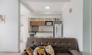 Ein modernes Wohnzimmer mit einer Couch und Kissen, eine Küche im Hintergrund und einer Uhr an der Wand.