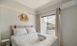 A neatly made bed with white linens, a headboard, and a rolled towel. A woven basket hangs on the wall, and there's a window with curtains. - Studio w/ BBQ Near the Airport - Rrc0206 (Florianópolis)