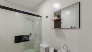 Un baño moderno con cabina de ducha de vidrio, inodoro, lavabo con espejo y un pequeño estante con una planta.