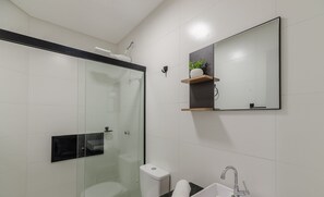A modern bathroom with a glass shower enclosure, a toilet, a sink with a mirror, and a small shelf with a plant. - Studio w/ BBQ Near the Airport - Rrc0206 (Florianópolis)