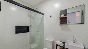 Moderni kylpyhuone, jossa on lasiseinällä varustettu suihku, wc, pesuallas ja peili, jonka yläpuolella on hylly.