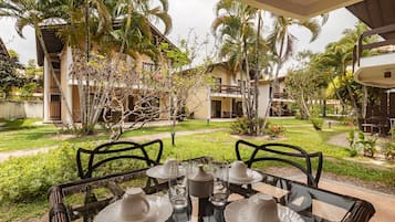 A table set for four with plates, glasses, and a teapot, overlooking a garden with palm trees and a building in the background.