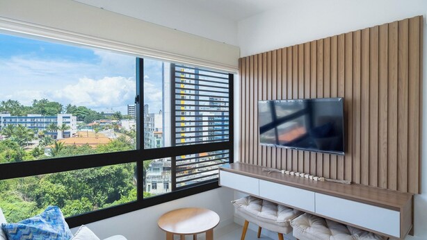 A modern living room with a large window, a flat-screen TV mounted on a wooden panel wall, a small round table, and a cushioned bench. - Apartment With Pool in Barra - Salvador Blb1004 (Salvador)