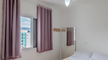 A bedroom with a bed, a window with curtains, a wall-mounted mirror, and a ceiling light.