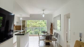 A modern kitchen with a flat-screen TV, a dining area with a glass table and chairs, and a view of greenery through large windows.
