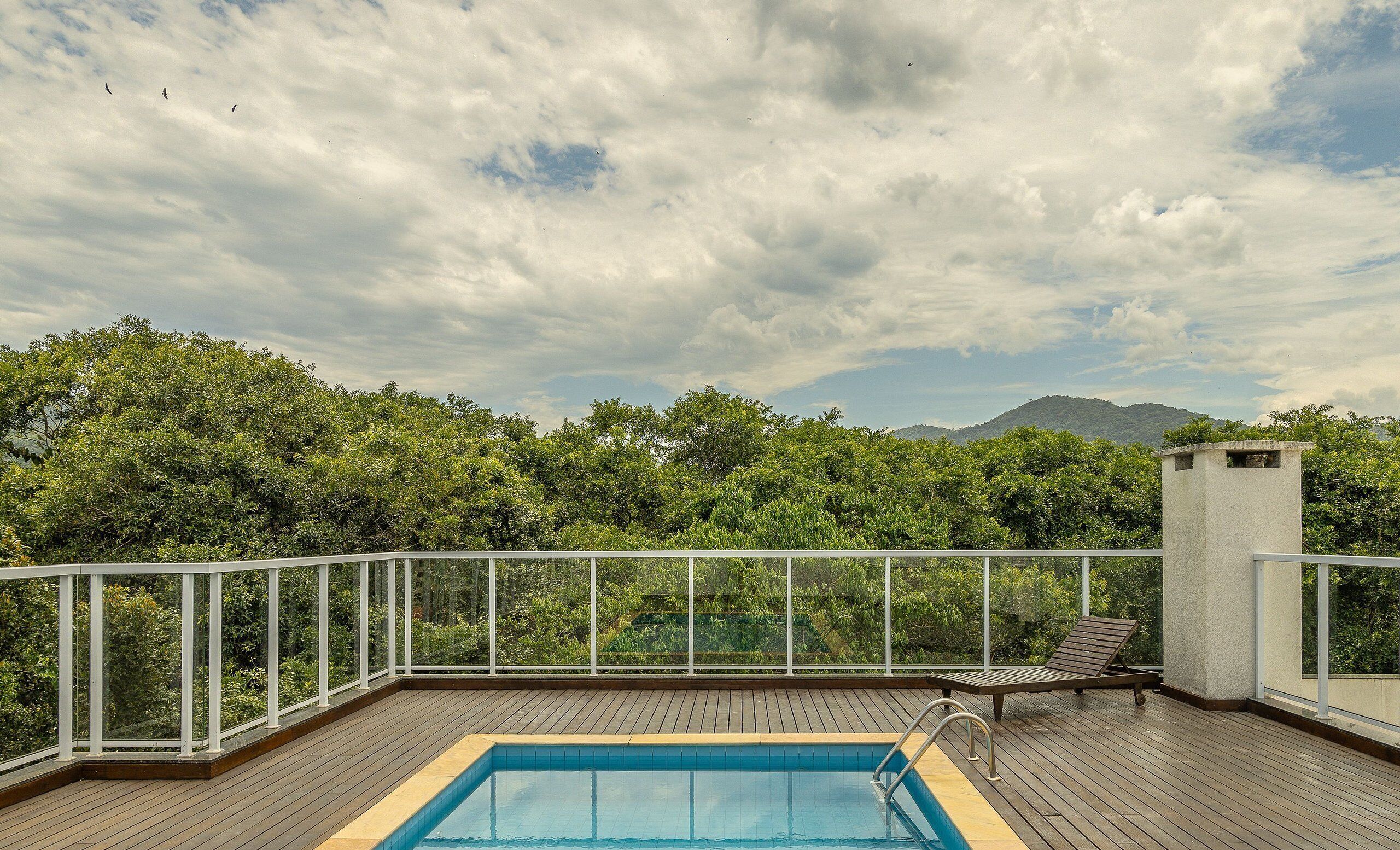 Une piscine sur le toit, dotée d’une terrasse en bois et entourée d’une balustrade blanche, offrant une vue imprenable sur un paysage verdoyant, avec des arbres et des montagnes à l’horizon.