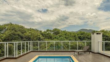 A rooftop pool with a wooden deck, surrounded by a white railing, and overlooking a lush green landscape with trees and distant mountains.