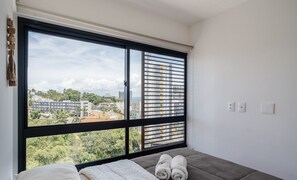 Ein Schlafzimmer mit großem Fenster, einem Bett und Blick auf Gebäude und Grünflächen.