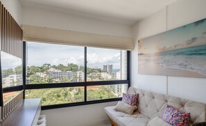 Ein modernes Wohnzimmer mit einem großen Fenster, einem beigefarbenen Sofa und einem farbenfrohen Gemälde vom Strand.