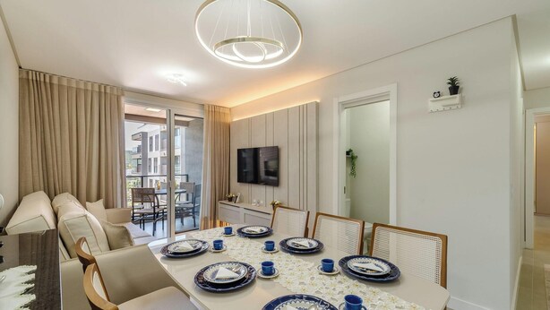 A dining area with a table set for six, a television mounted on the wall, and a view of the outside balcony. - Apart w/ Barbecue in Jurere Internacional Vlo0504 (Florianópolis)