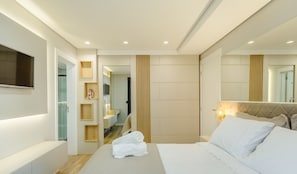 Quarto de hotel moderno, com uma cama, uma televisão e um banheiro visível por uma porta aberta.
