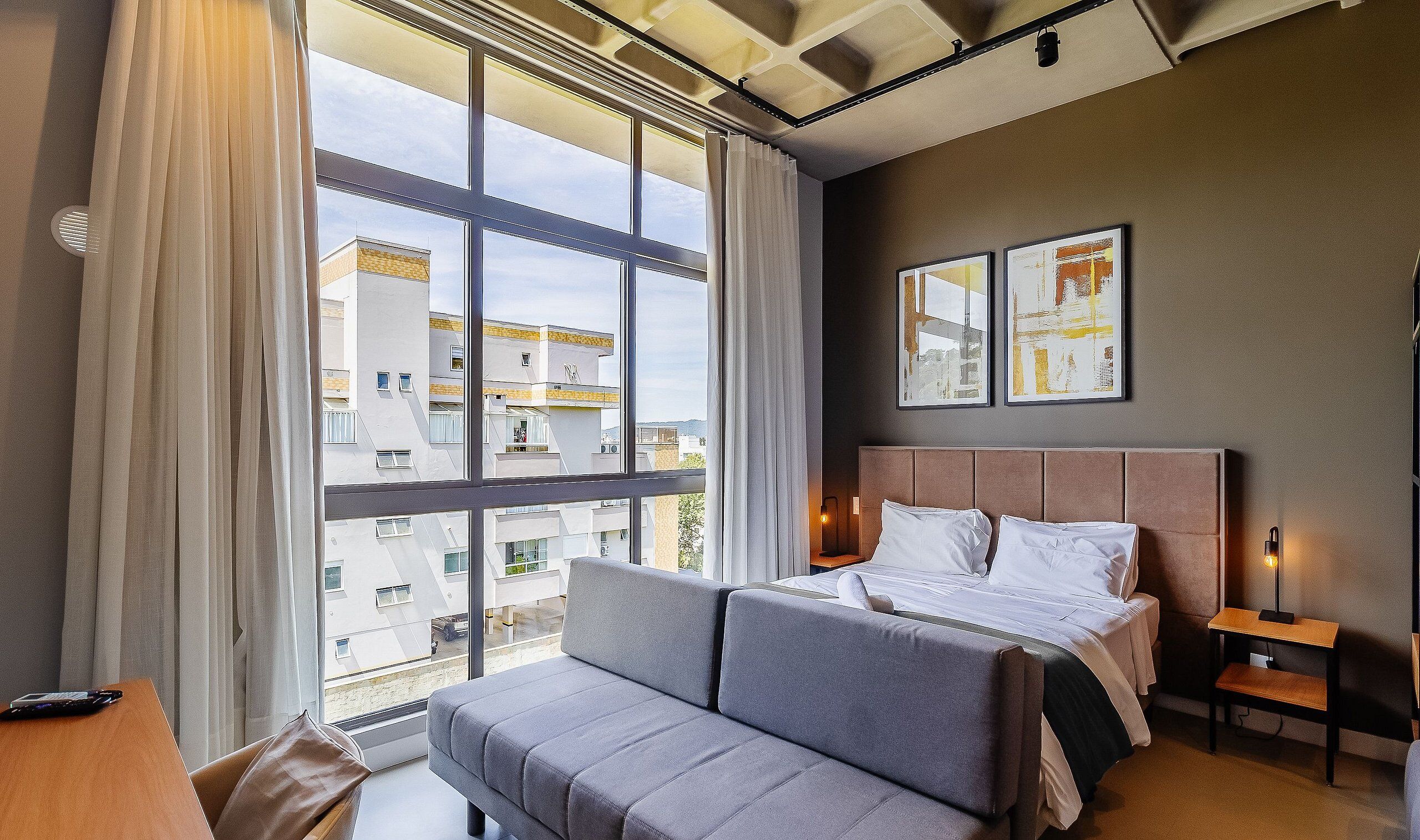 A hotel room with a large window, a bed, a sofa, and a bedside table.