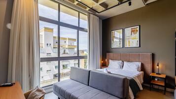 A hotel room with a large window, a bed, a sofa, and a bedside table.