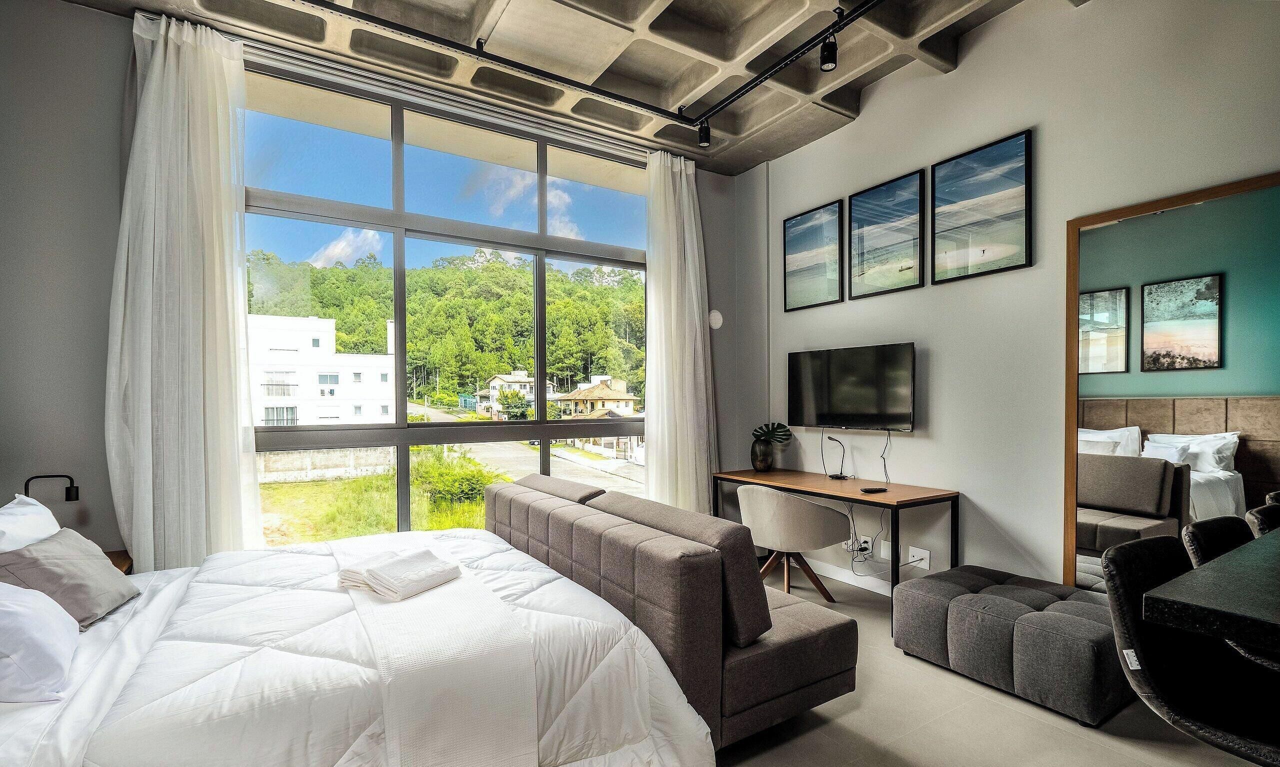 Habitación de hotel con cama, sofá, escritorio, televisor y ventanas amplias con vistas a edificios y árboles.