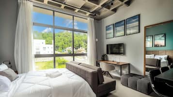 Habitación de hotel con cama, sofá, escritorio, televisor y ventanas amplias con vistas a edificios y árboles.
