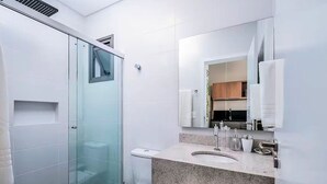 A modern bathroom with a glass shower enclosure, a sink with a granite countertop, and a large mirror reflecting a kitchen area. - Amazing Studio 750m From Canas Beach Tps105 (Florianópolis)