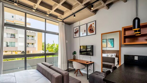 A modern hotel room with a large window, a bed, a desk, a TV, and artwork on the walls. - Amazing Studio 750m From Canas Beach Tps105 (Florianópolis)