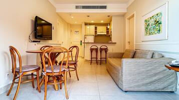 A dining area with wooden chairs and a table, a sofa, a television, and a kitchen in the background.