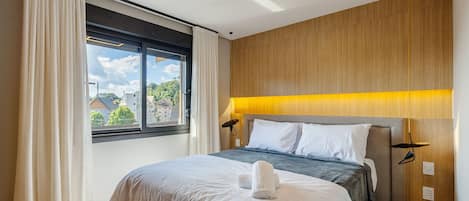 A hotel room with a bed, a window with curtains, a wooden headboard, and a bedside lamp.