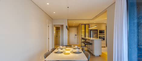 Un espace de repas moderne avec une longue table dressée pour quatre personnes, une cuisine en arrière-plan et une grande fenêtre offrant une belle vue.