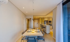 A modern dining area with a long table set for four, a kitchen in the background, and a large window with frosted glass. - Apt in the Penthouse With Barbecue! - Dlh0501 (Gramado)