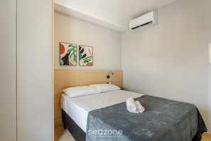 Ein ordentlich gemachtes Bett mit grauer Decke und aufgerollten weißen Handtüchern, ein Holz-Kopfteil und zwei abstrakte Gemälde an der Wand.
