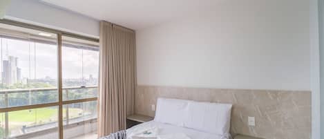 Una habitación de hotel con un ventanal grande, una cama con sábanas blancas, una manta gris y una pared de mármol.