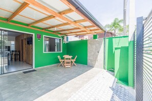 Um alpendre coberto com mesa e cadeiras de madeira, uma parede verde e uma porta de vidro deslizante que dá para um espaço interno.