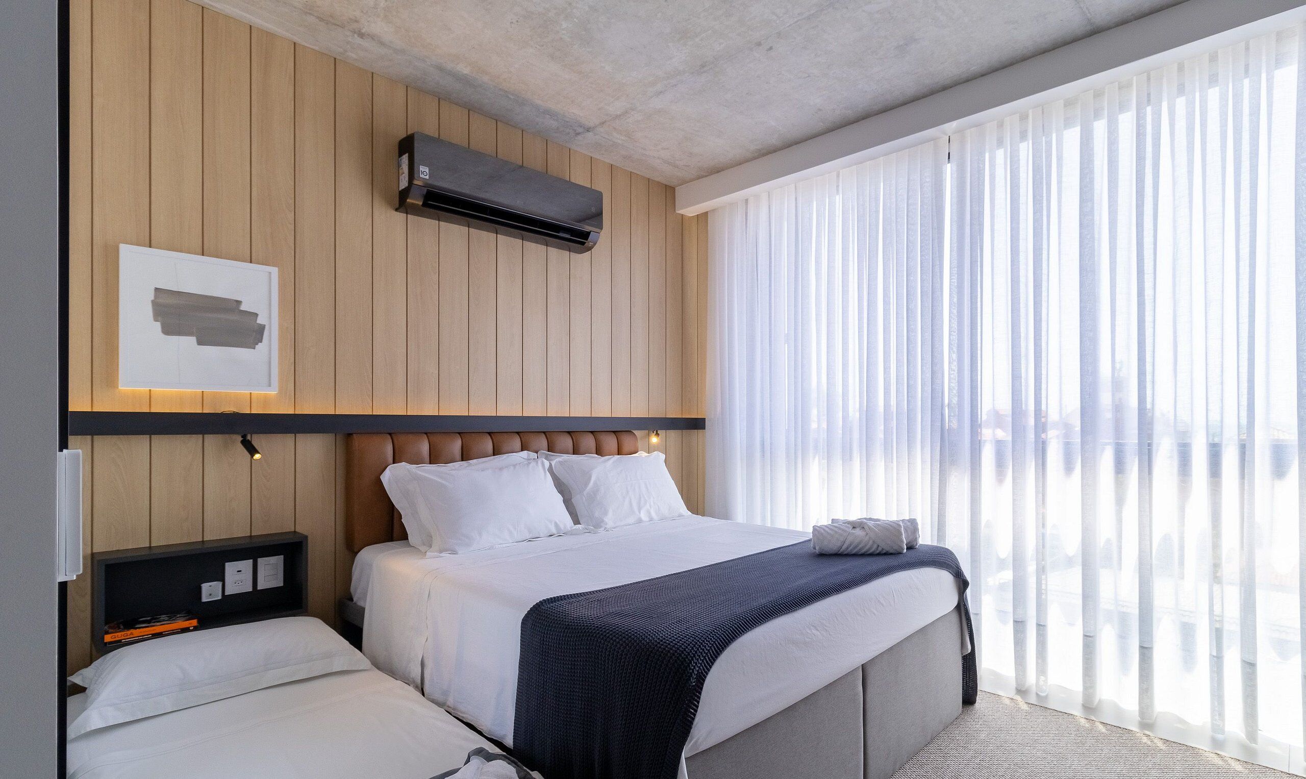 A modern hotel room with a bed, bedside tables, an air conditioning unit, and a large window with sheer curtains.