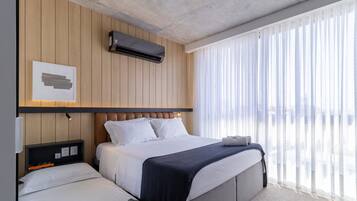 A modern hotel room with a bed, bedside tables, an air conditioning unit, and a large window with sheer curtains.
