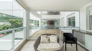 Un balcone con una sedia in vimini e cuscini, una sedia nera e vista su un edificio e vegetazione.