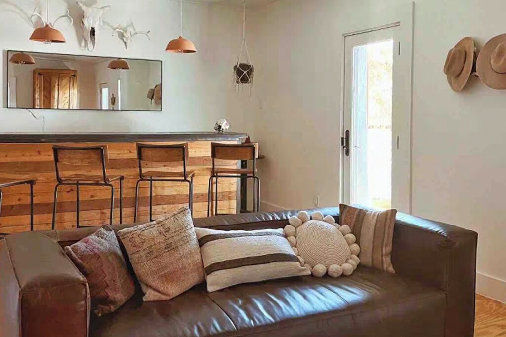 Una sala de estar con un sofá de cuero, una mesa de comedor de madera y sillas. Hay un espejo en la pared y una puerta con sombreros colgados.