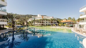 Une piscine d’un complexe hôtelier, avec une eau d’un bleu limpide, entourée de palmiers et de bâtiments modernes.