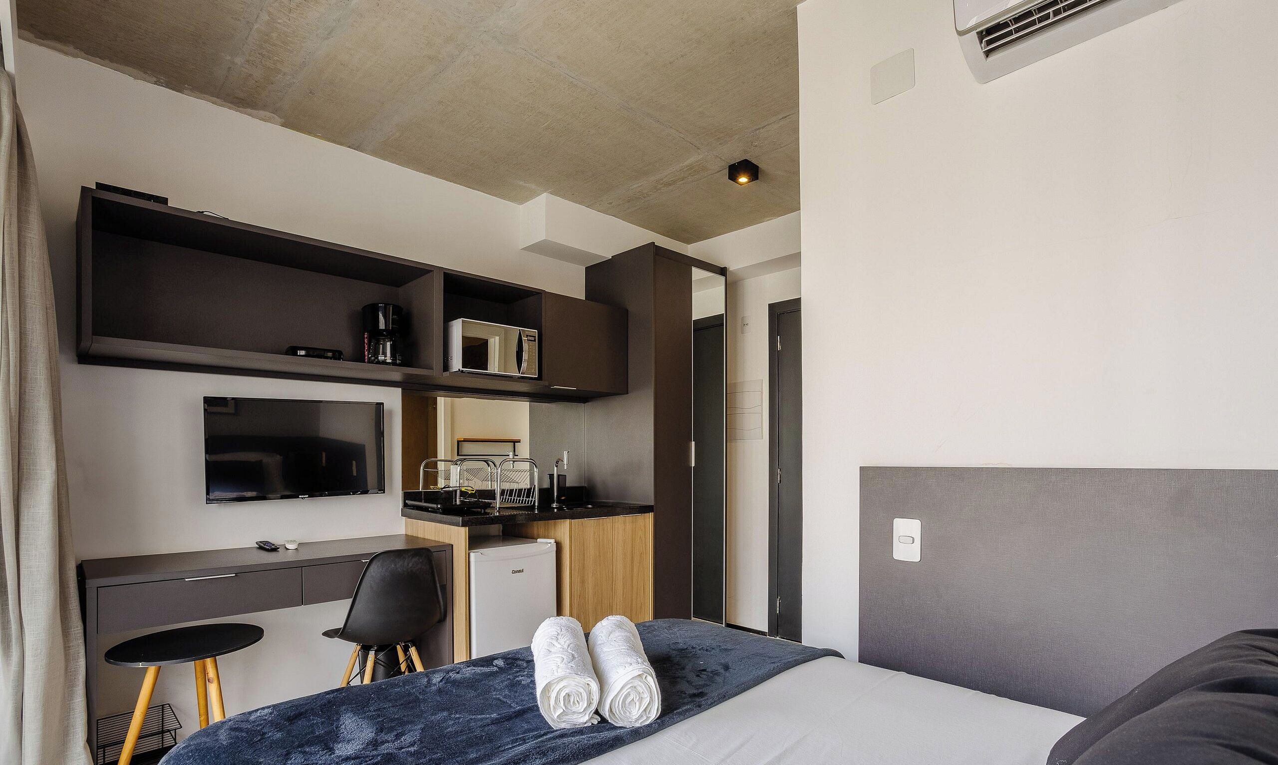 A compact hotel room with a bed, a desk, a chair, a small kitchenette, and a television.