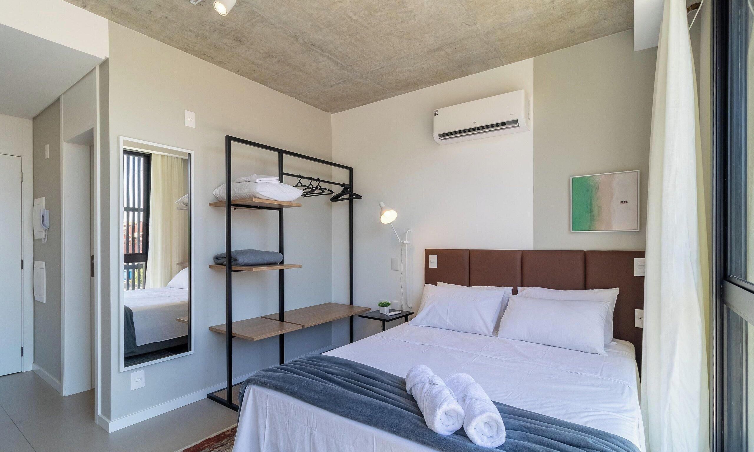 Une chambre d’hôtel comprenant un lit, une armoire avec des étagères, une table de chevet, un climatiseur fixé au mur et une fenêtre avec des rideaux.