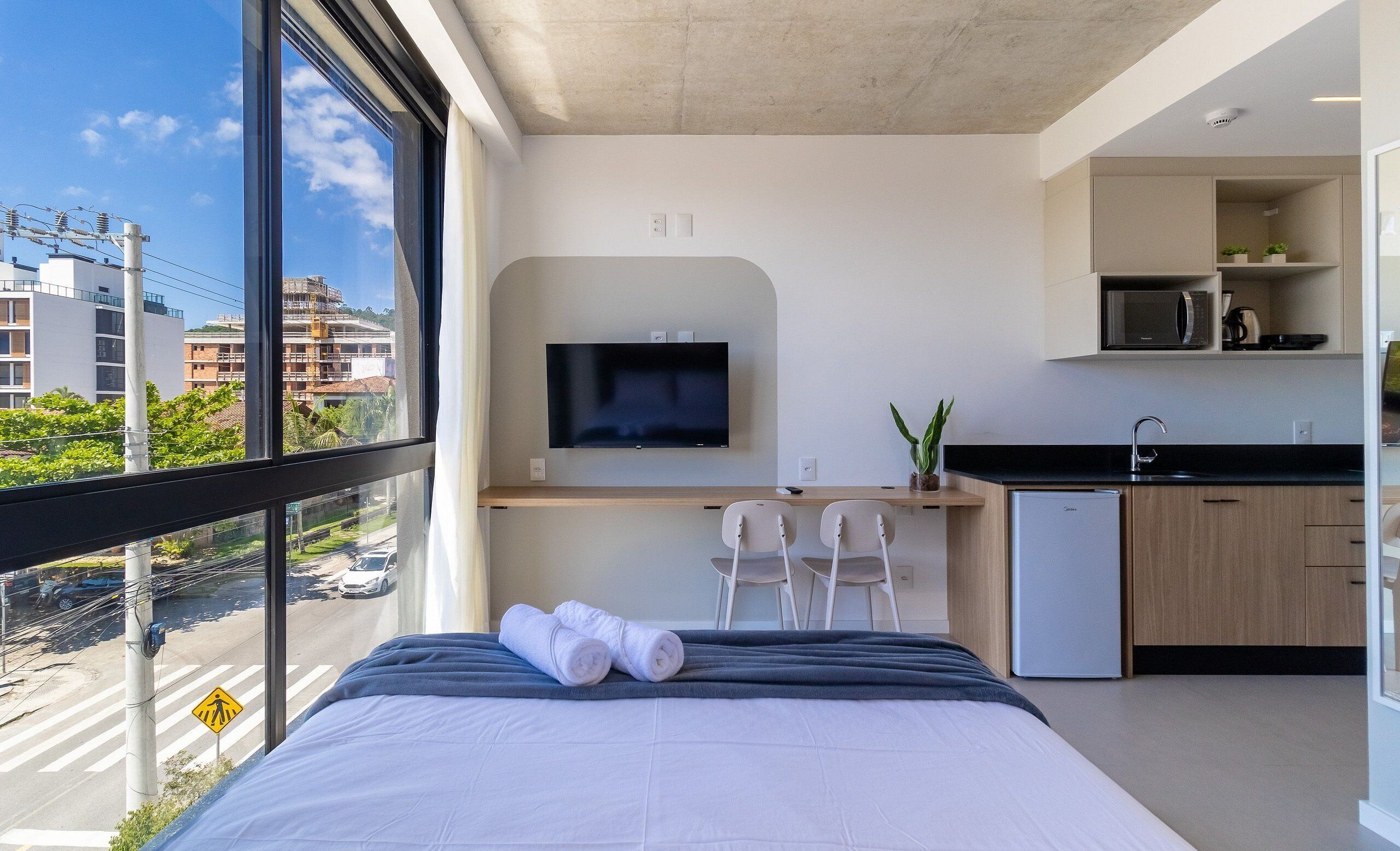 A modern hotel room with a bed, a desk with a TV, a small kitchenette, and a view of the city.