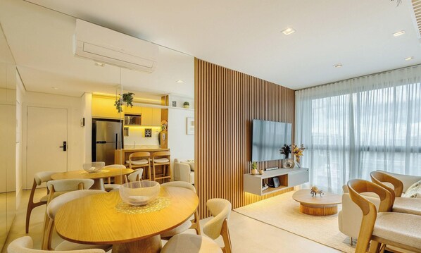 A modern living room with a round wooden dining table, cushioned chairs, a TV, and a kitchen area in the background. - Modern and Complete Stay in Brasilia - Epk0903 (Guará)