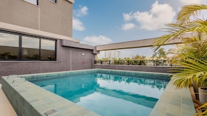 Una piscina en la azotea con borde de azulejos, rodeada por un edificio con grandes ventanales y vistas a un paisaje urbano.