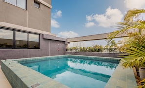 Una piscina en la azotea con borde de azulejos, rodeada por un edificio con grandes ventanales y vistas a un paisaje urbano.