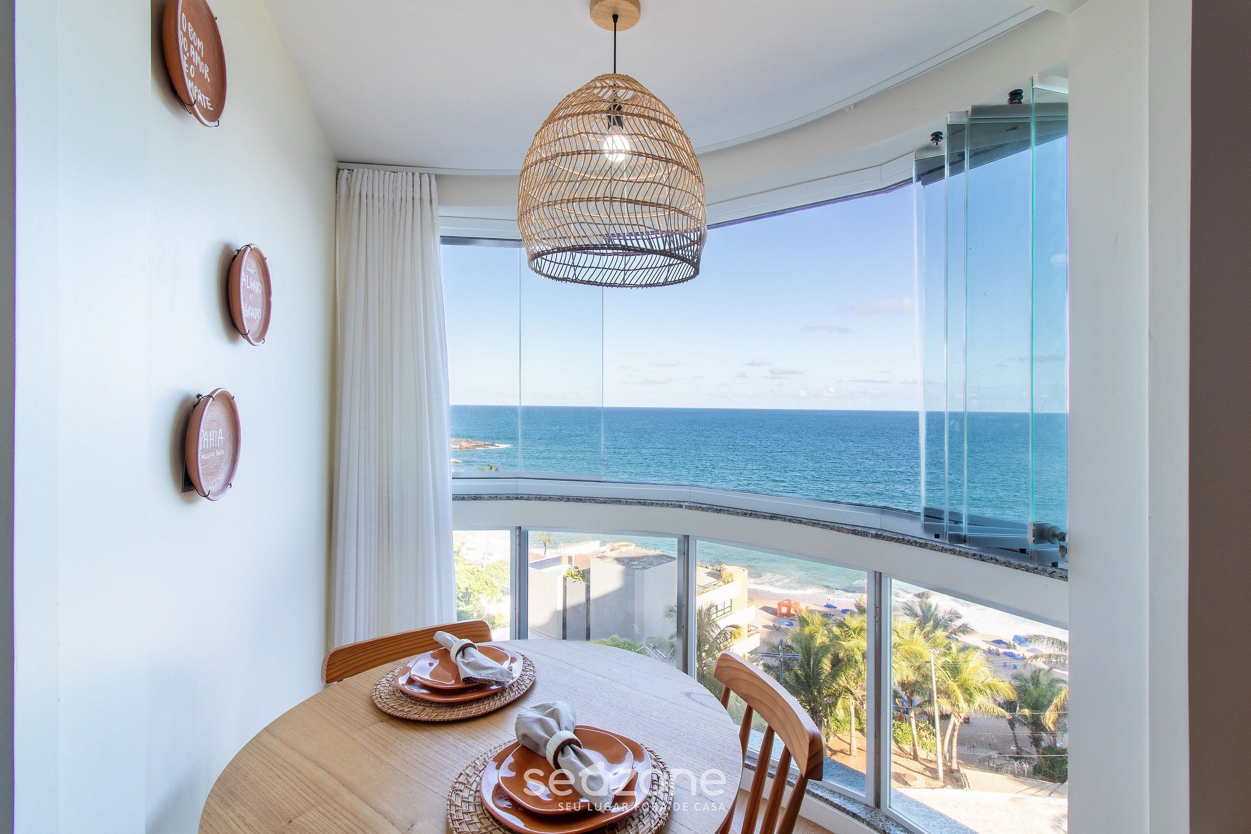 A dining area with a view of the sea, a wicker pendant light, and a round table with plates and a napkin.