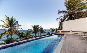 En swimmingpool med udsigt over havet, omgivet af palmer og en hvid bygning.
