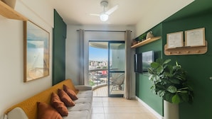 A modern living room with a sofa, a ceiling fan, a large window with a view of the city, and a green accent wall. - Apt in Cond. w/ Pool, Steps fr the Beach Esn0702 (Salvador)