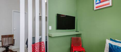 Una habitación con pared verde, un televisor montado, dos sillas rojas, una silla de madera y una puerta que da a otra habitación.
